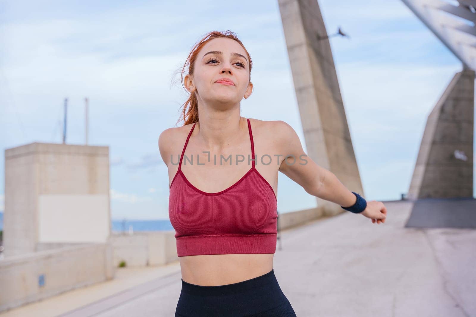 Fit red-haired woman wearing sportswear stretching her arms behind her back in an outdoor urban setting. She prepares for exercise under a bright sky, radiating energy and confidence.