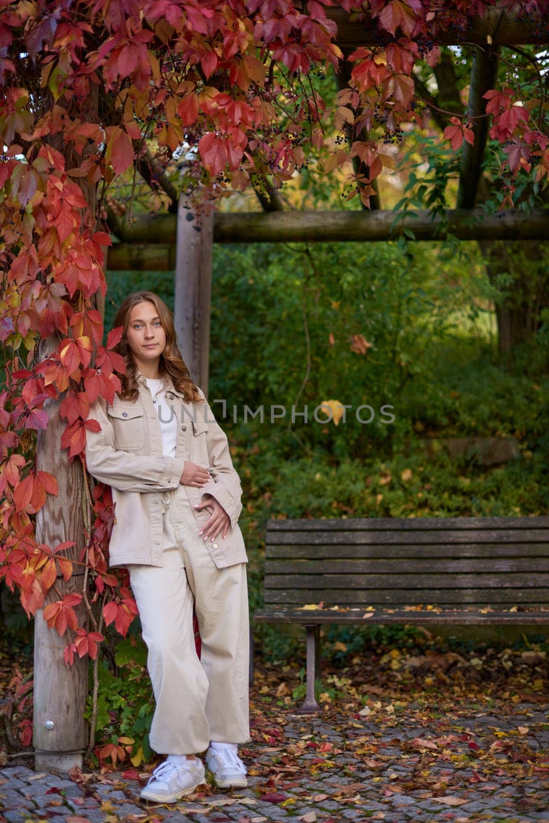 A young woman with wavy light brown hair and a cheerful smile is leaning against a wooden pillar wrapped in red autumn leaves. She is dressed in a beige casual outfit, including a stylish denim jacket and matching pants, with a white top underneath. The background features a scenic garden pergola with wooden beams covered in lush green and red foliage, creating a natural and cozy atmosphere. The warm lighting and soft depth of field enhance the autumn aesthetic, making this image perfect for seasonal fashion, lifestyle blogs, outdoor portraiture, and autumn-themed marketing campaigns. Smiling young woman in beige casual outfit leaning on wooden pillar with red autumn leaves in scenic garden pergola – warm and cozy fall portrait with natural background