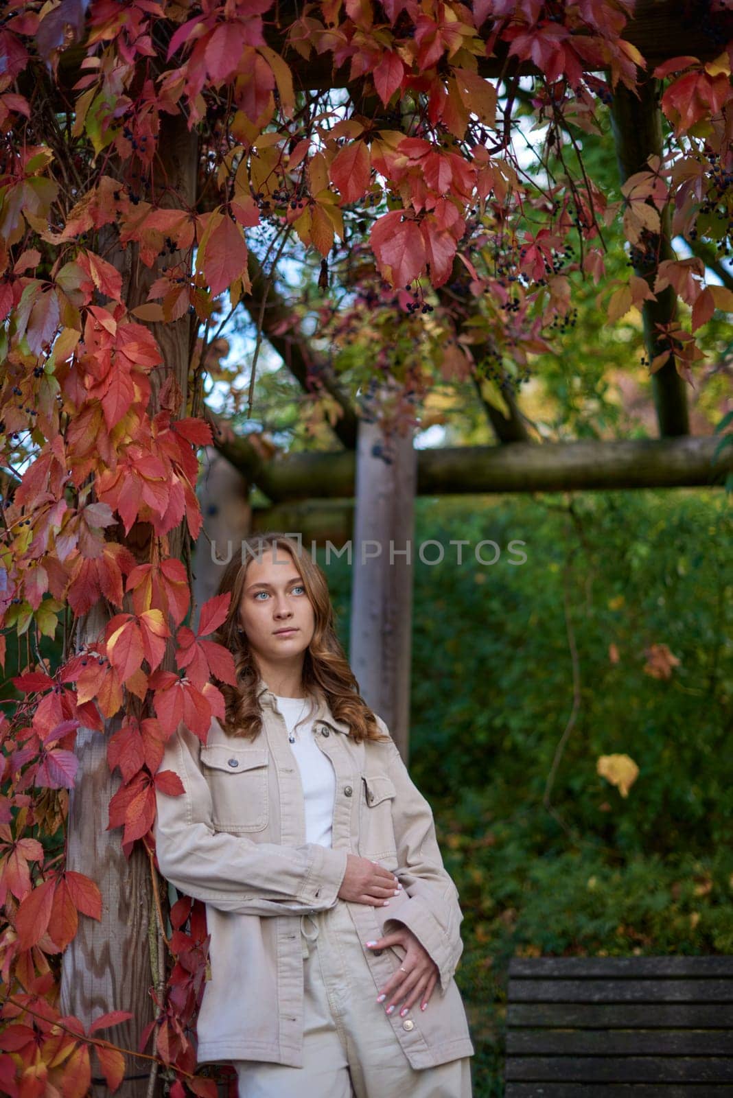 Capture the essence of autumn with this charming portrait of a young woman embracing the season's warmth and beauty. The subject, dressed in a casual beige outfit, leans comfortably against a wooden pillar of a garden pergola, her smile radiating contentment and joy. Vibrant red autumn leaves frame the scene, creating a stunning natural backdrop that epitomizes fall's rich palette. The rustic pergola adds a touch of countryside charm, while the overall composition exudes a cozy, inviting atmosphere. This image is perfect for autumn-themed marketing campaigns, lifestyle blogs, fashion editorials, and travel publications, showcasing the beauty of outdoor living and seasonal transitions.