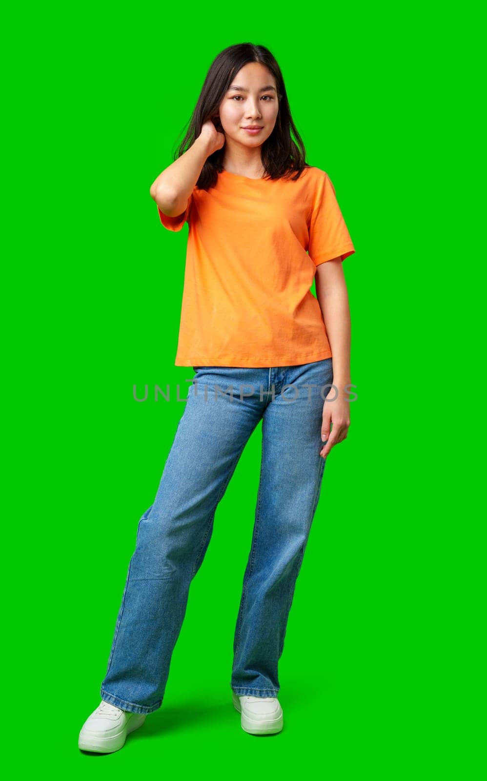 An individual stands in an orange shirt and blue jeans, showcasing a relaxed pose on a vivid green backdrop. The setting emphasizes her outfit and confidence.