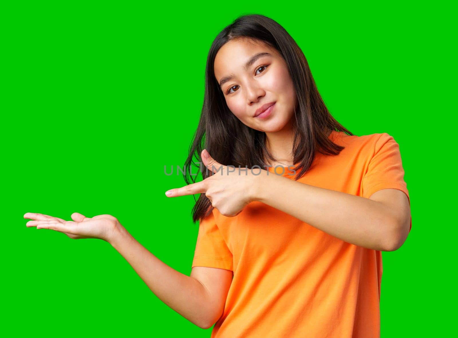A young woman wearing an orange shirt stands confidently on a bright green background. She gestures with her right hand, pointing to an invisible space as if inviting attention to something important.