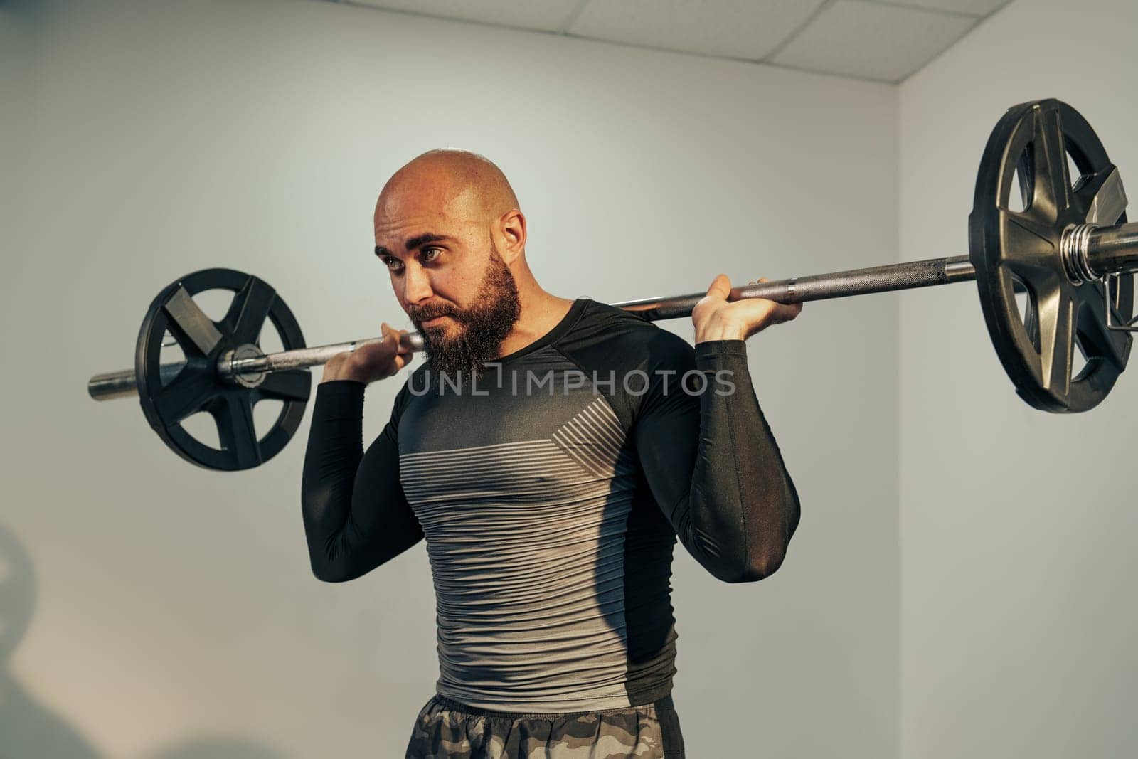 A man with a beard and bald head performs a barbell squat in a gym, showcasing his strength and focus. The workout occurs in a well-lit space early in the day.