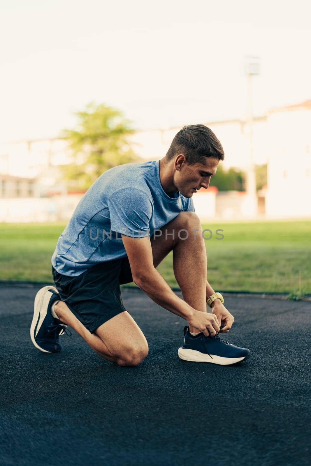 A young man kneels on a track field, tying his running shoes as he prepares for a workout. The soft evening light casts a warm glow over the scene, enhancing his focus and determination.