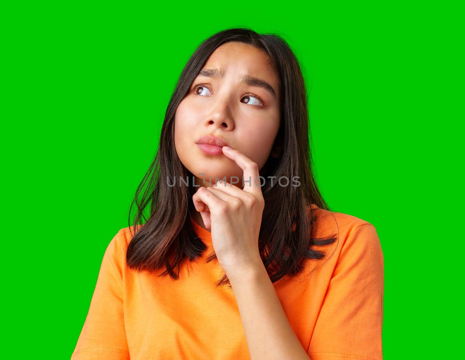 A young woman with long dark hair poses thoughtfully against a vibrant green backdrop. She rests her finger on her chin, indicating deep thinking or curiosity.