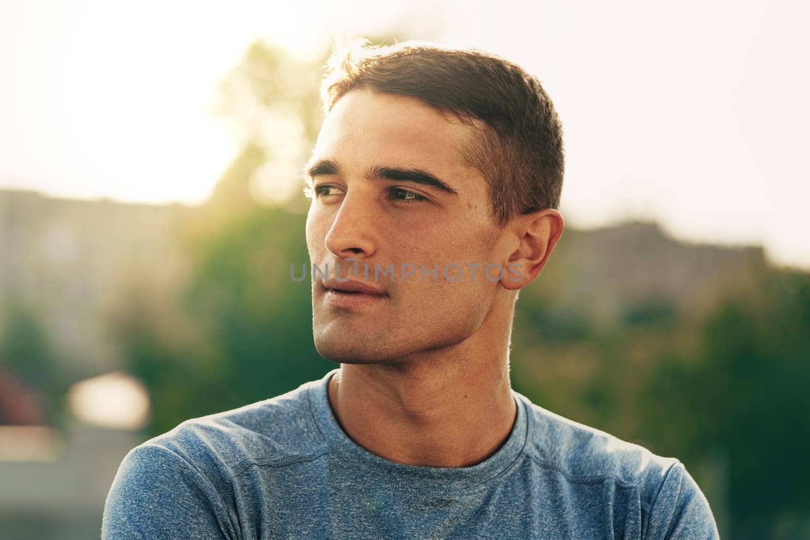 A young man stands outdoors, wearing a blue shirt. The warm light of the golden hour highlights his features as he gazes into the distance, lost in thought.