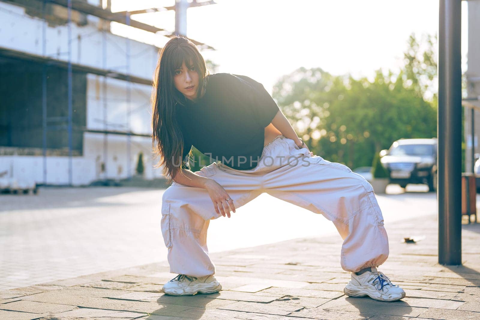 A dancer showcases her skills on the street in an urban setting. She wears casual attire and exhibits an energetic pose. The sun shines brightly, creating a dynamic atmosphere.