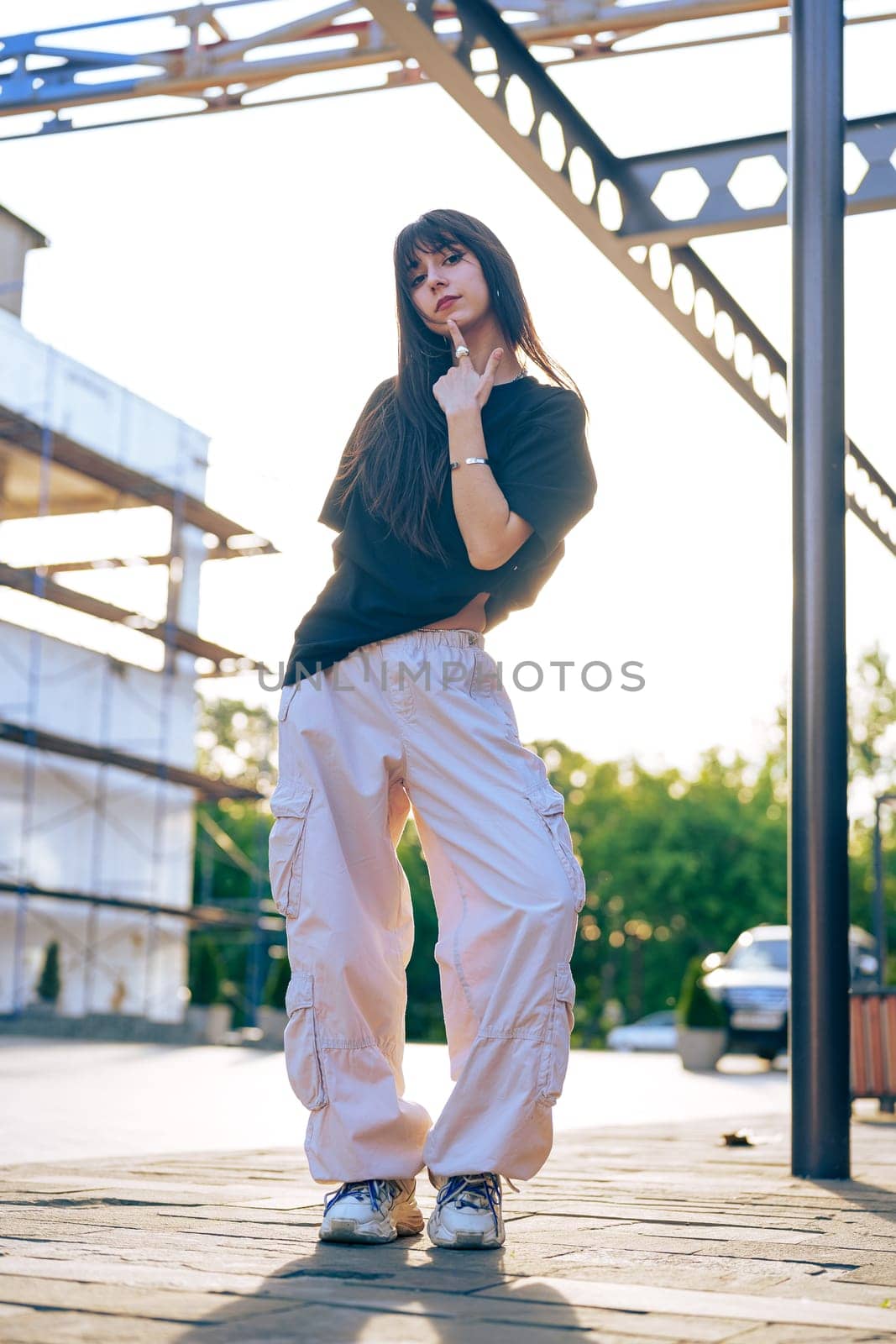 A woman stands confidently with one hand on her chin, wearing casual streetwear. The warm sunlight illuminates a contemporary background of construction, capturing a vibrant moment.