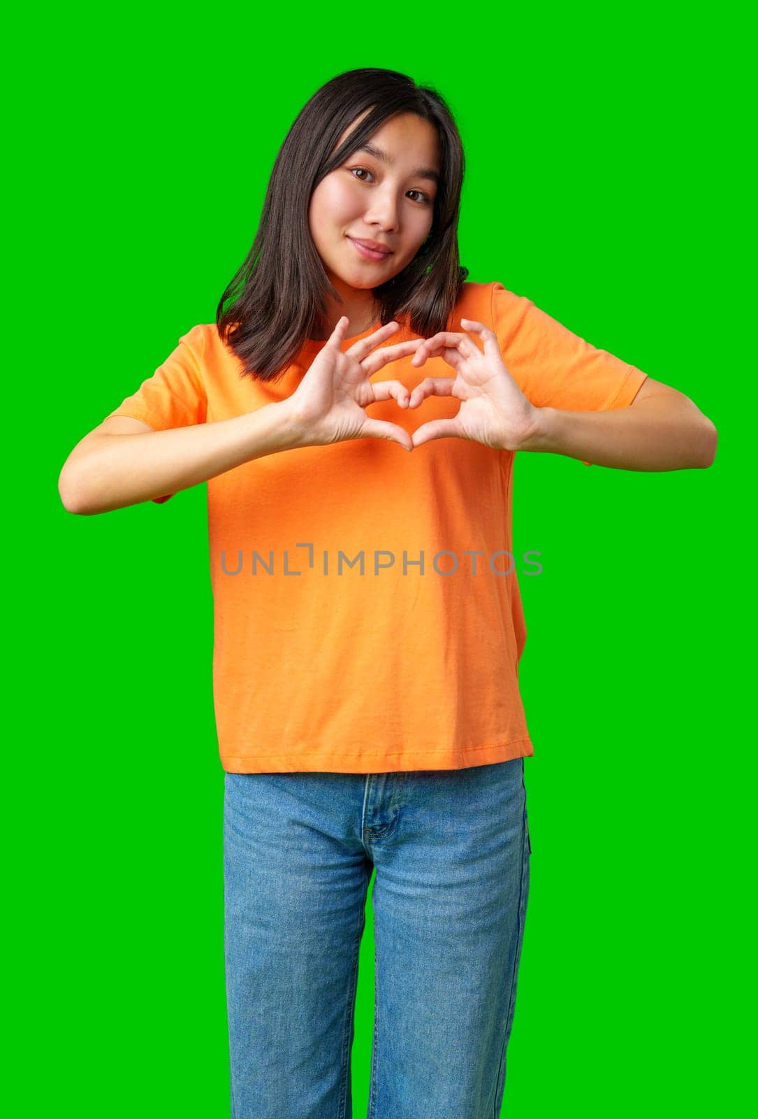 A young individual wearing an orange shirt forms a heart shape with their hands, smiling playfully in front of a vivid green backdrop. The atmosphere is lively and cheerful.