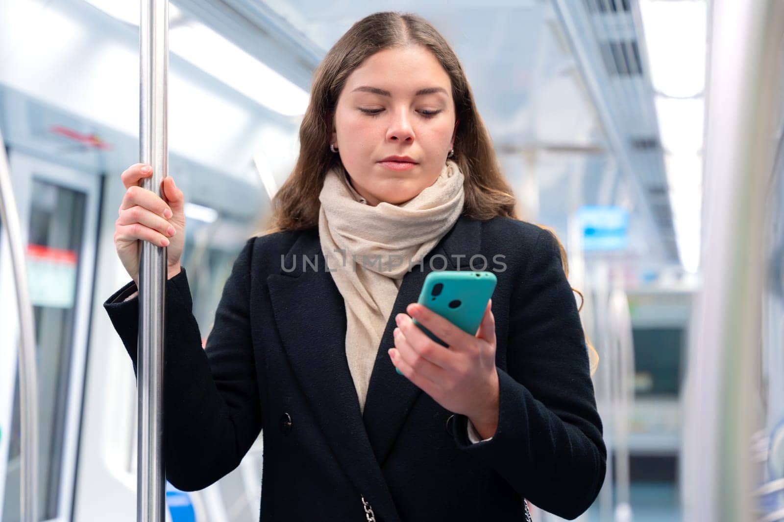 Young woman in a black coat and beige scarf standing inside a modern subway train, holding a smartphone and gripping a metal pole. She appears focused while commuting, carrying a stylish white purse.