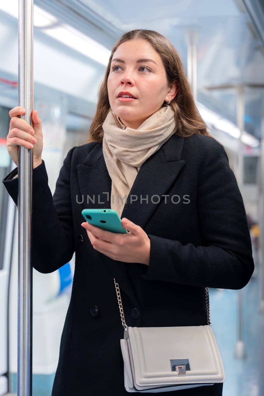 Young woman in a black coat and beige scarf standing inside a modern subway train, holding a smartphone and gripping a metal pole. She appears focused while commuting, carrying a stylish white purse.