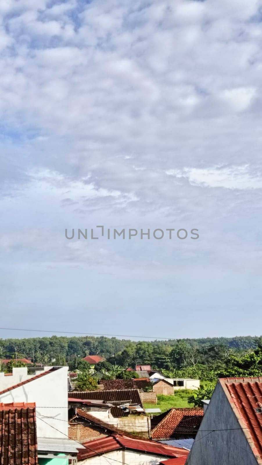 Tranquil view of a serene village with red-tiled roofs, surrounded by greenery, under a partly cloudy sky. Captures peaceful rural living and a connection to nature.