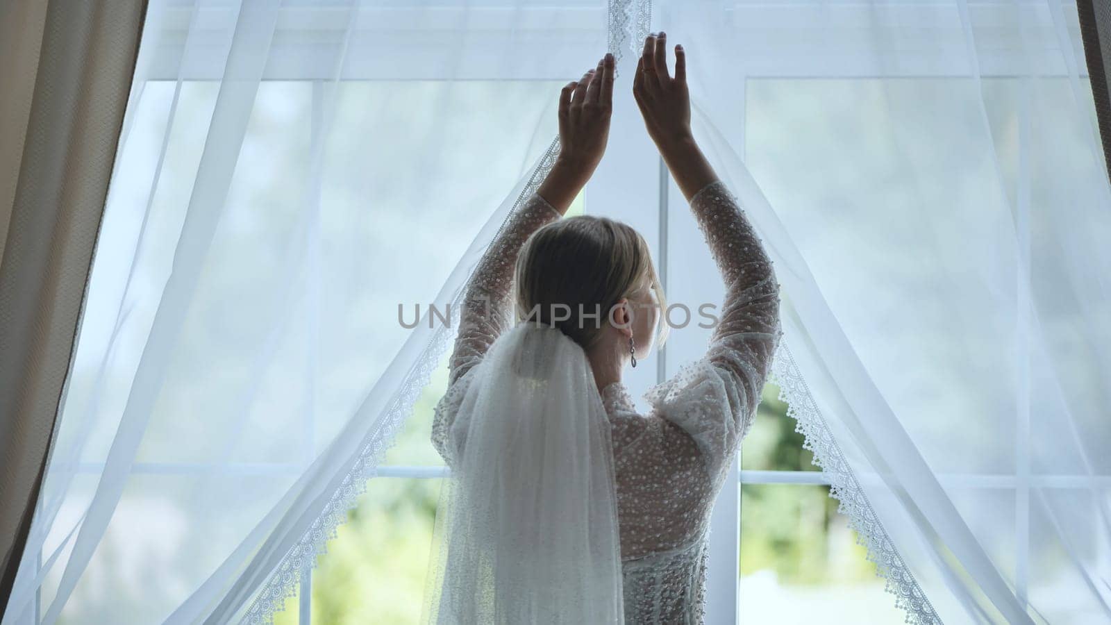 Bride in flowing wedding dress revealing sunlit window, soft veil trailing, capturing serene morning wedding preparation moment