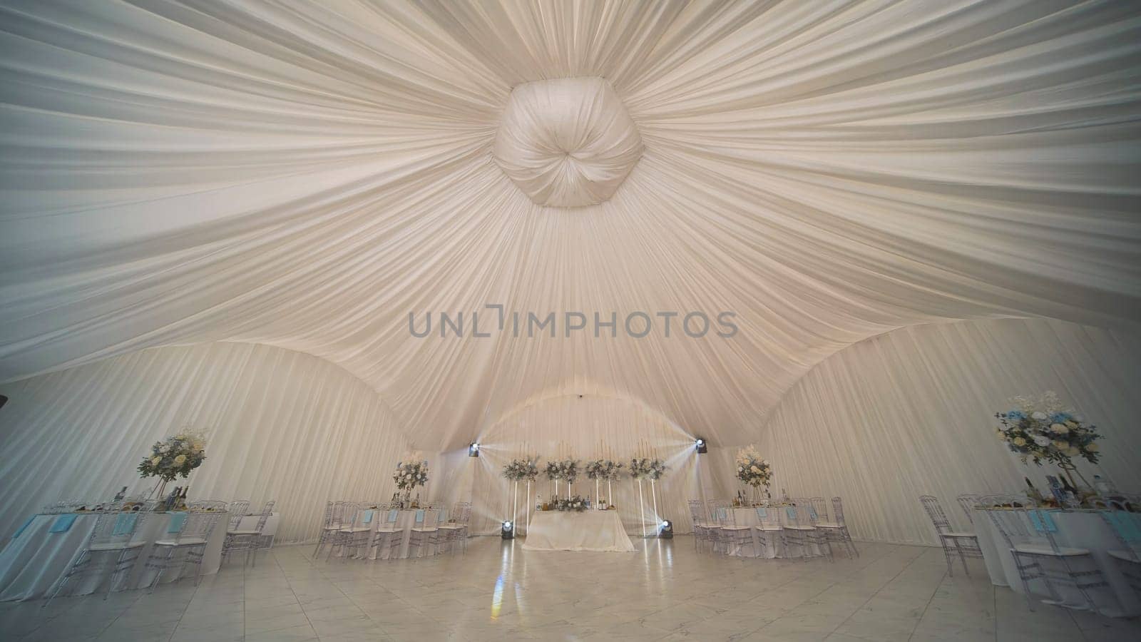 Elegant tent decorated with white ceiling drapery and flower arrangements, set up for a wedding reception, creating a romantic and luxurious atmosphere