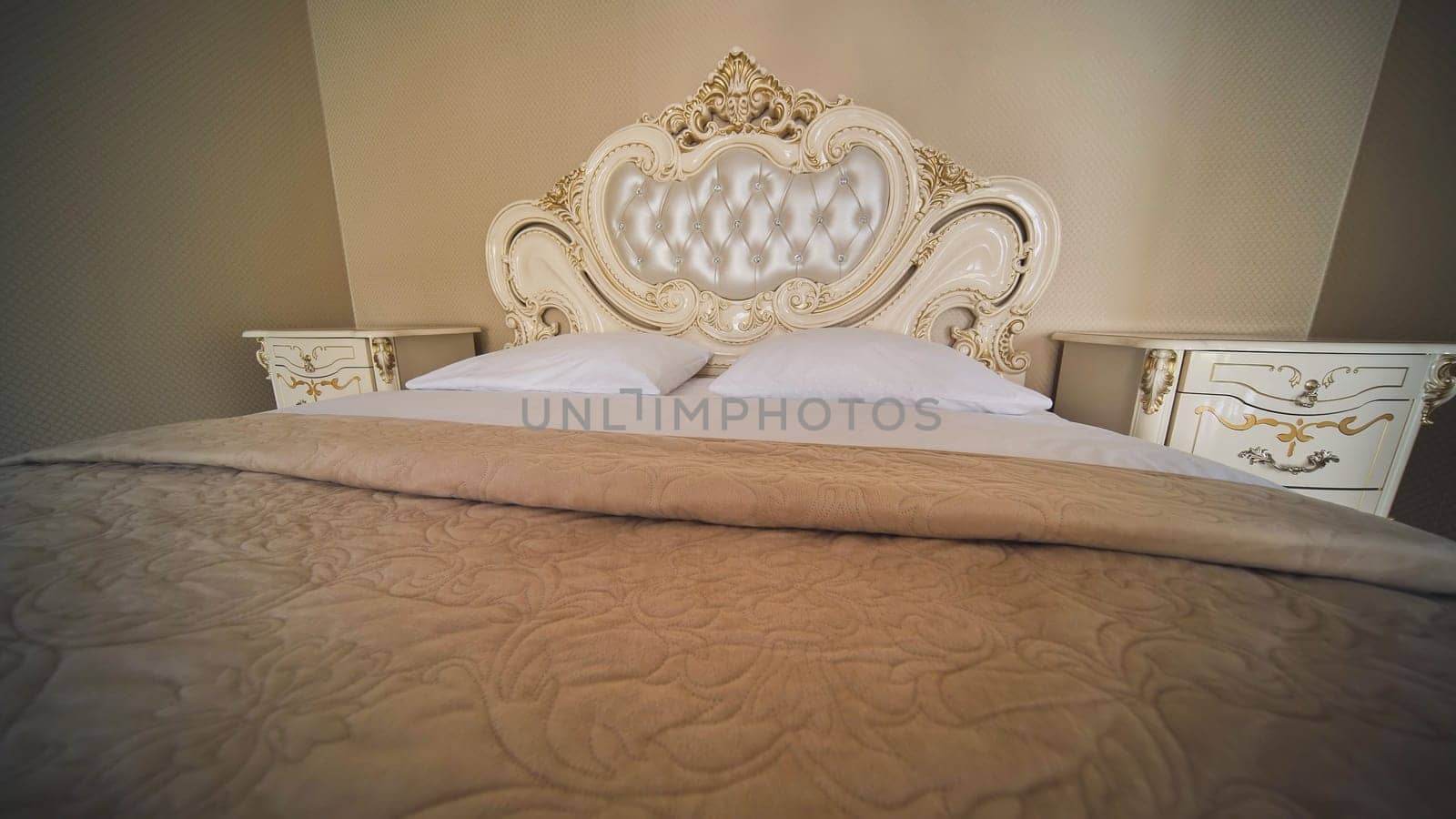 Luxurious beige king size bed with ornate headboard, bedside tables, and soft blanket, creating romantic atmosphere for wedding night or honeymoon