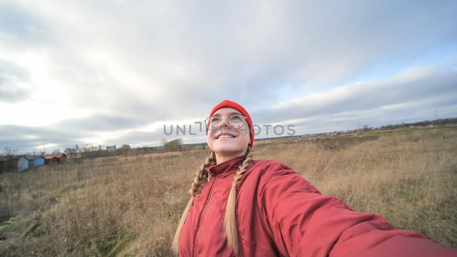 Smiling young woman with red winter clothes and glasses taking selfie in a countryside field, enjoying cloudy sky