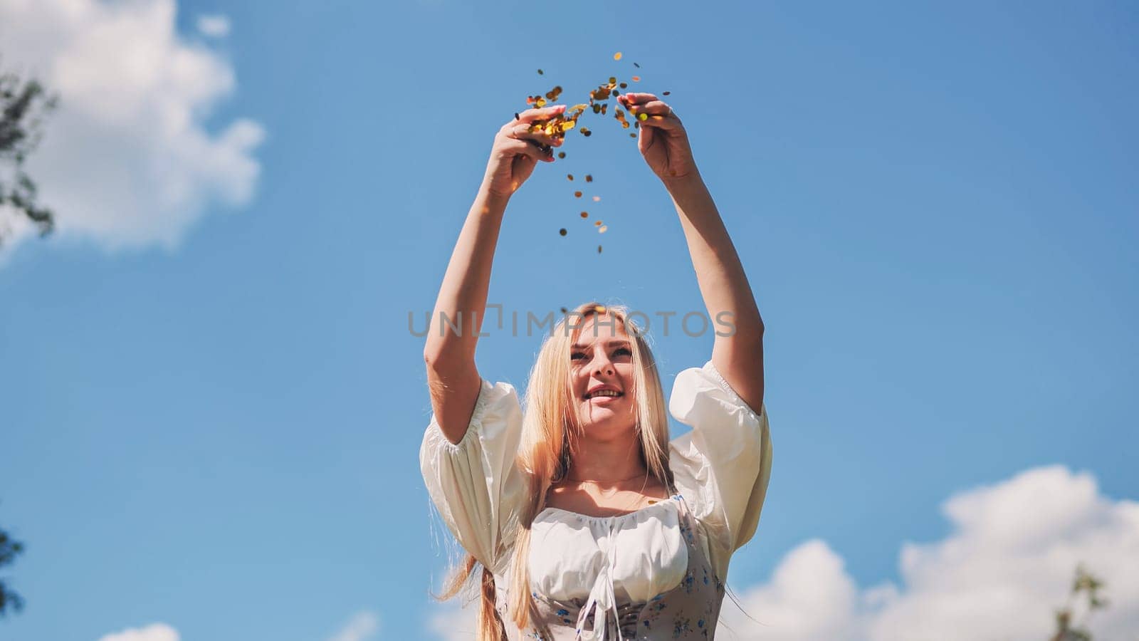 Slavic woman throwing gold confetti in the air, celebrating against a bright blue sky with fluffy white clouds