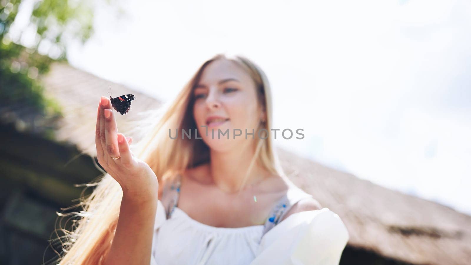 Blonde woman resting outdoors, delicate butterfly perching gently on palm amid lush green landscape, embodying serene connection with nature