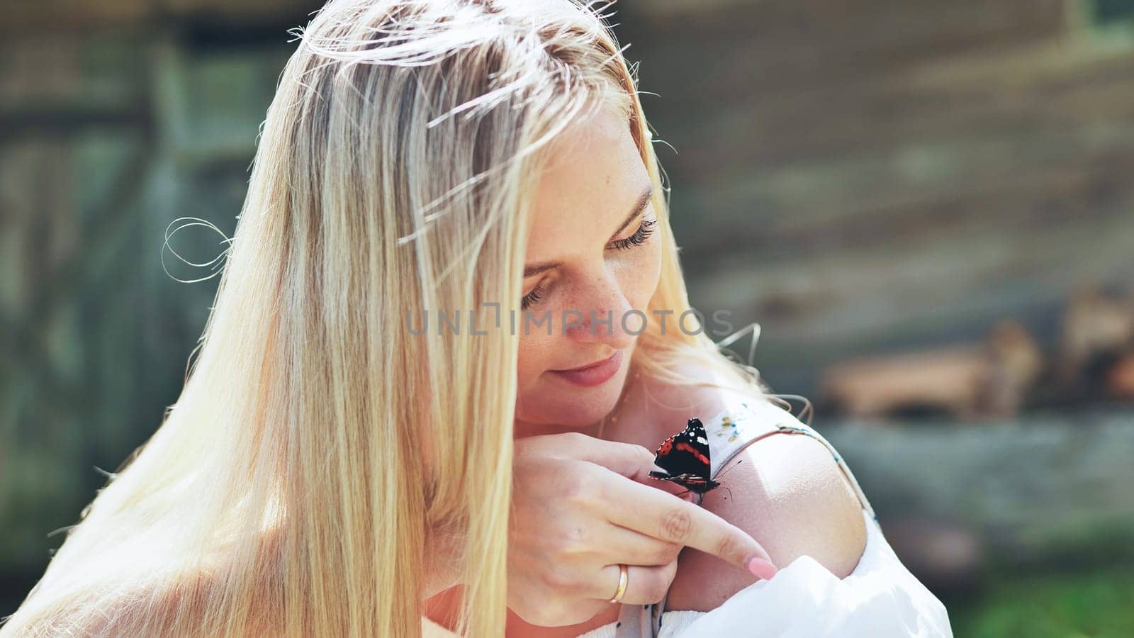 Blond woman with freckles gently touching butterfly standing on her shoulder with index finger