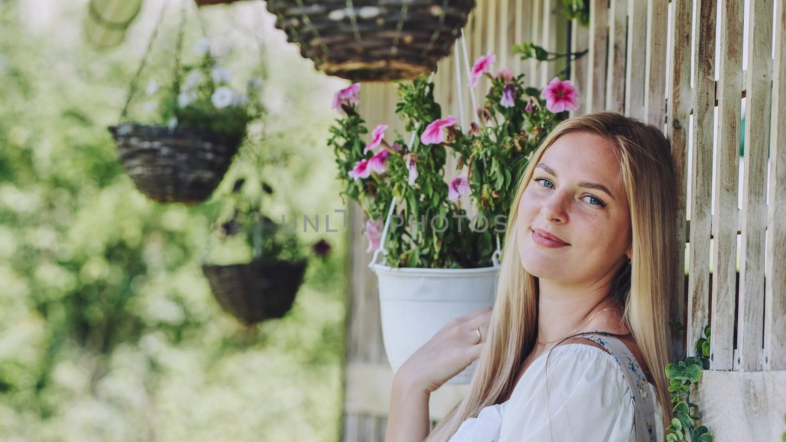 Blonde woman resting among hanging flower baskets, basking in serene garden landscape with gentle sunlight