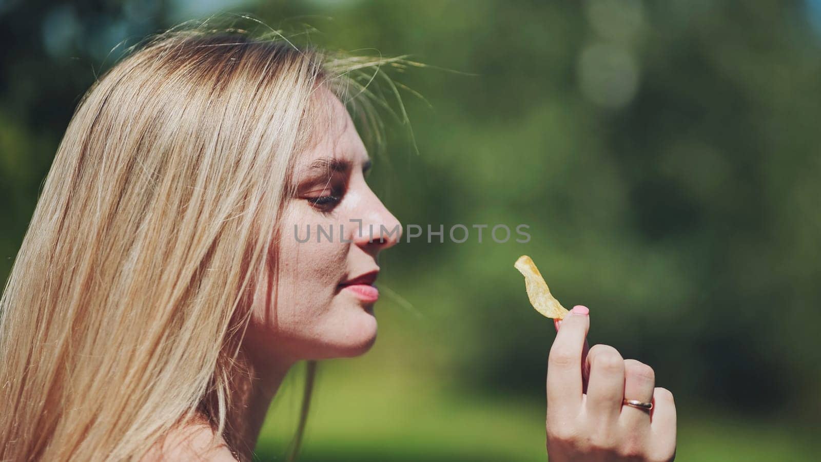 Profile of a blonde woman holding a potato chip between her fingers outdoors on a summer day