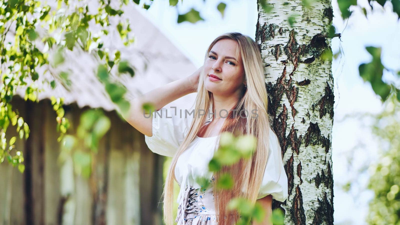 Blond woman in traditional slavic attire standing near birch, rural setting capturing cultural heritage and pastoral scenery