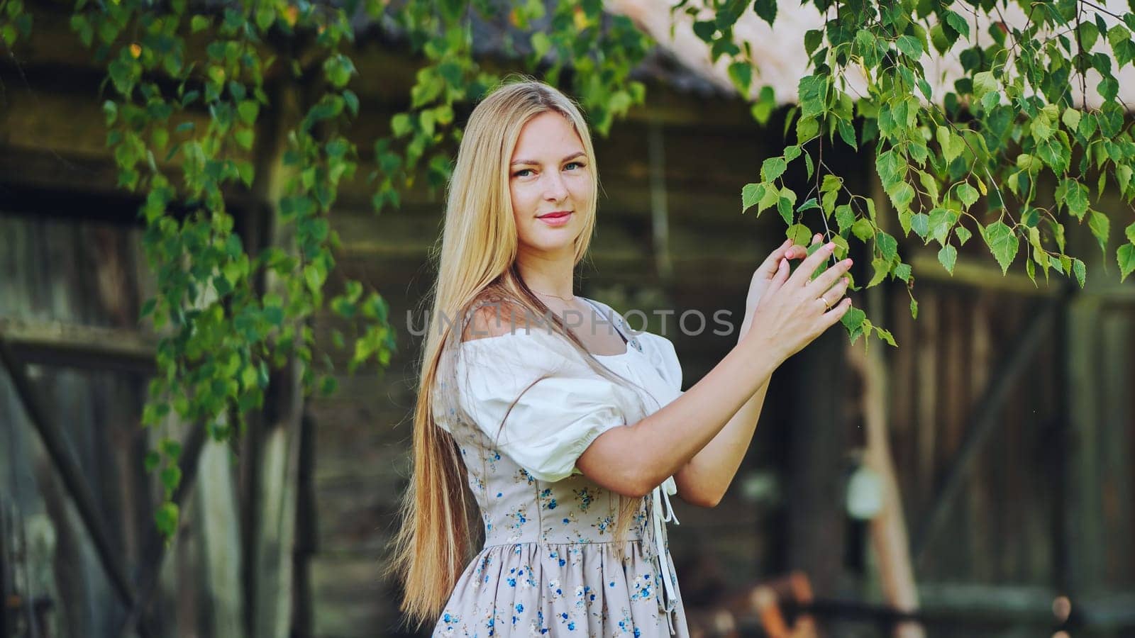 Young blonde woman wearing traditional slavic dress gently touching birch leaves in a peaceful countryside setting