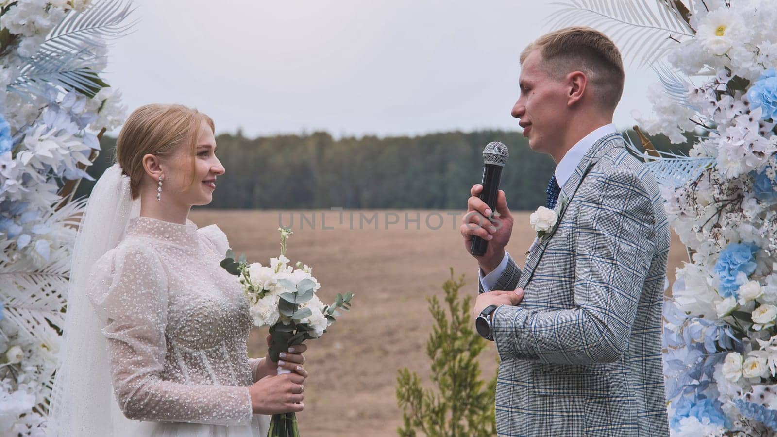 Loving groom reading handwritten vows, speaking into microphone, gazing adoringly at bride during rustic outdoor wedding ceremony
