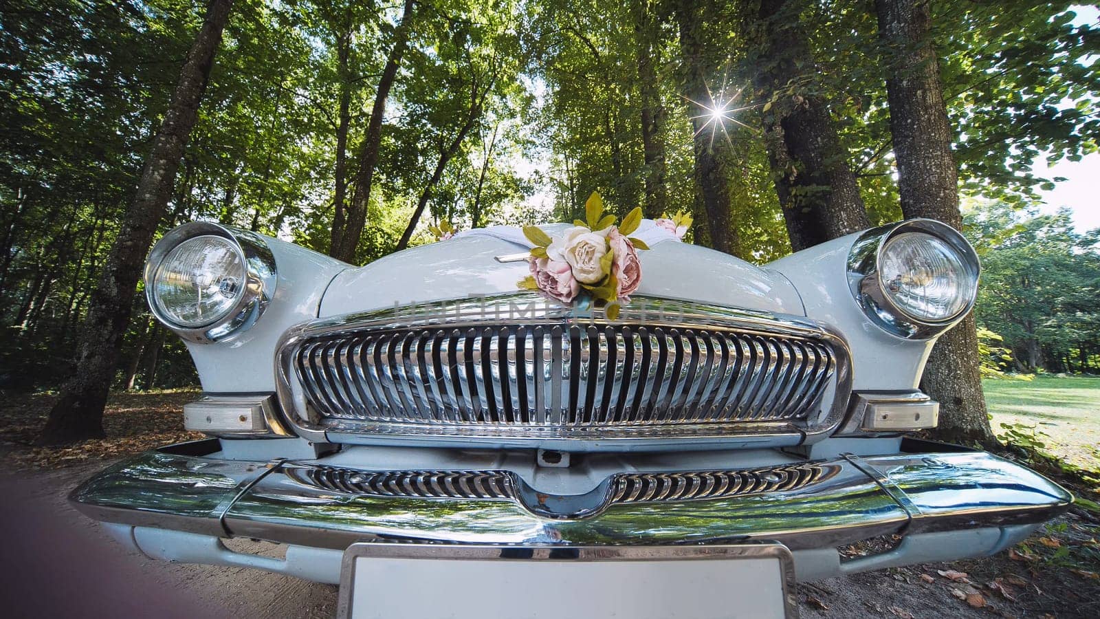 Vintage white car adorned with floral bouquet, parked near trees at wedding venue, symbolizing romantic celebration