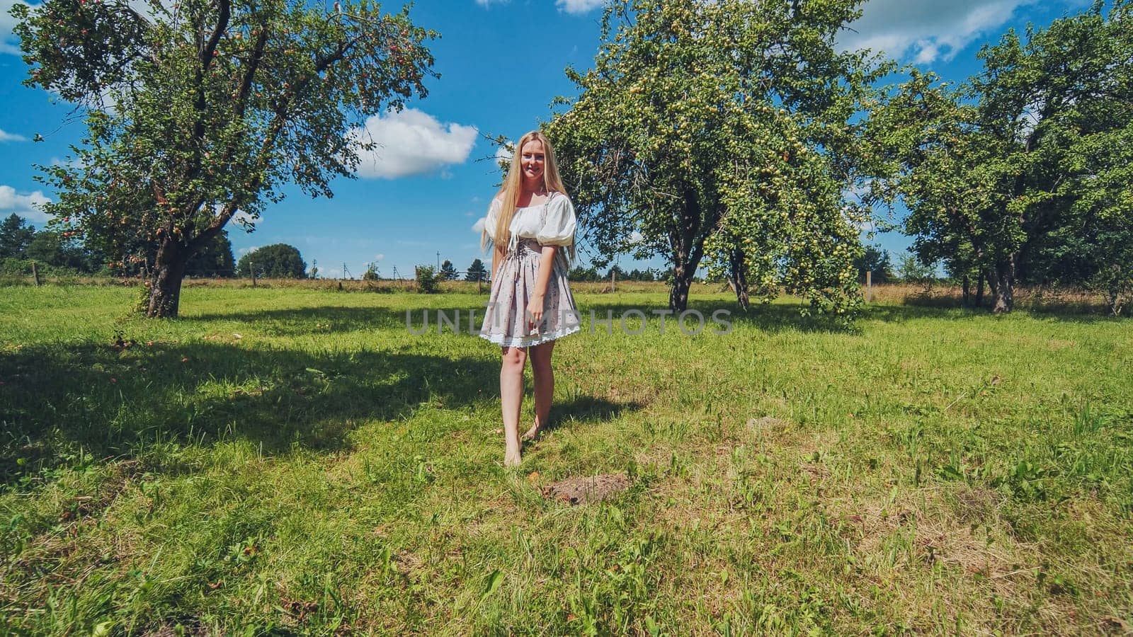 Blonde woman wearing traditional slavic clothes walking barefoot in a green meadow under a summer blue sky