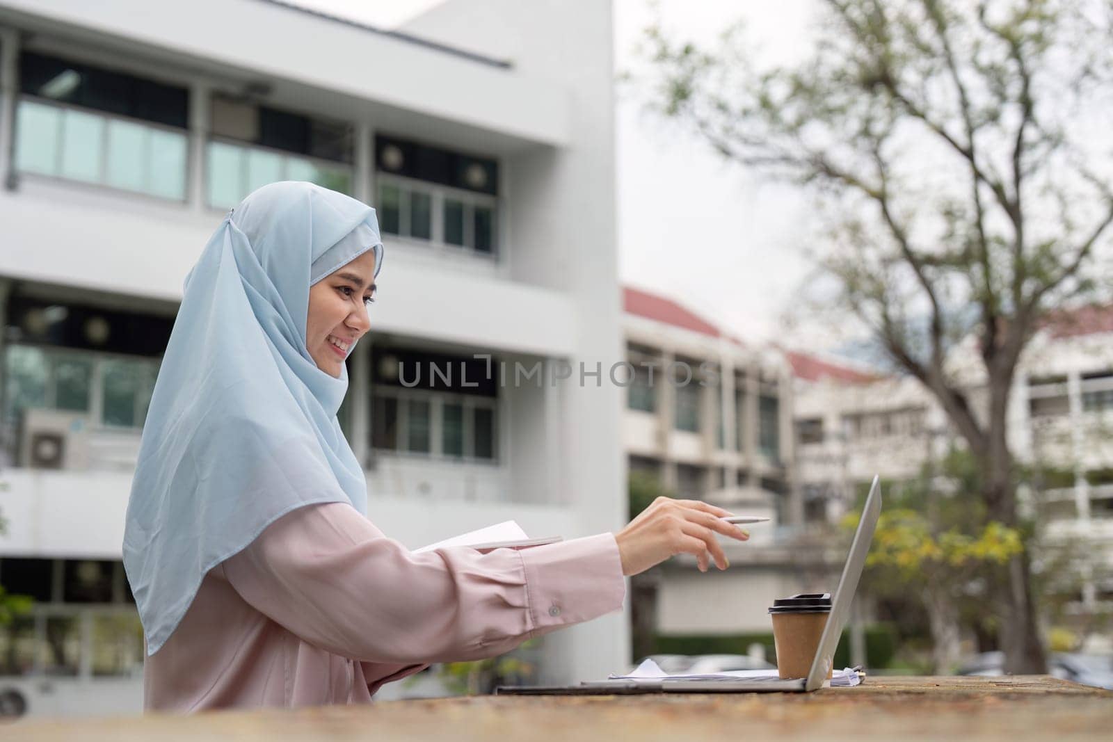 A cheerful Muslim businesswoman engaged in her work on a laptop, enjoying a coffee break in a vibrant outdoor setting.