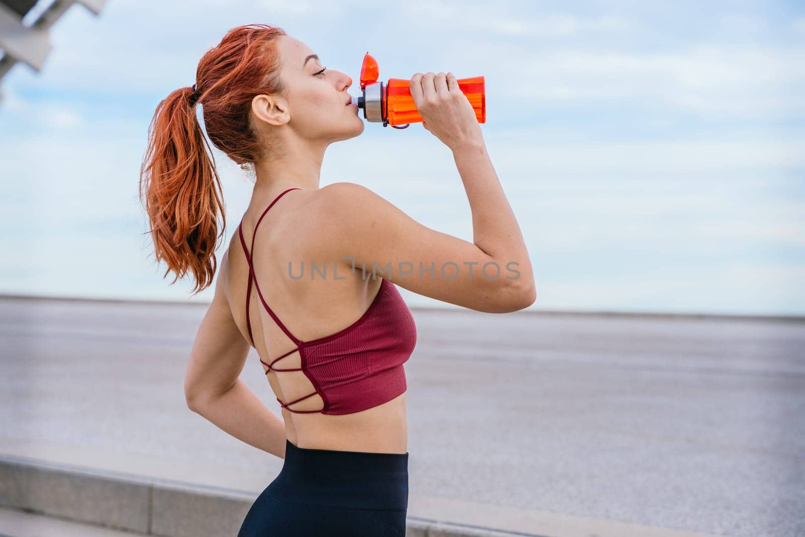 Athletic red-haired woman wearing a stylish sports bra and leggings, drinking water after exercising. She stands outdoors in an urban environment under a bright blue sky, staying hydrated and energized.