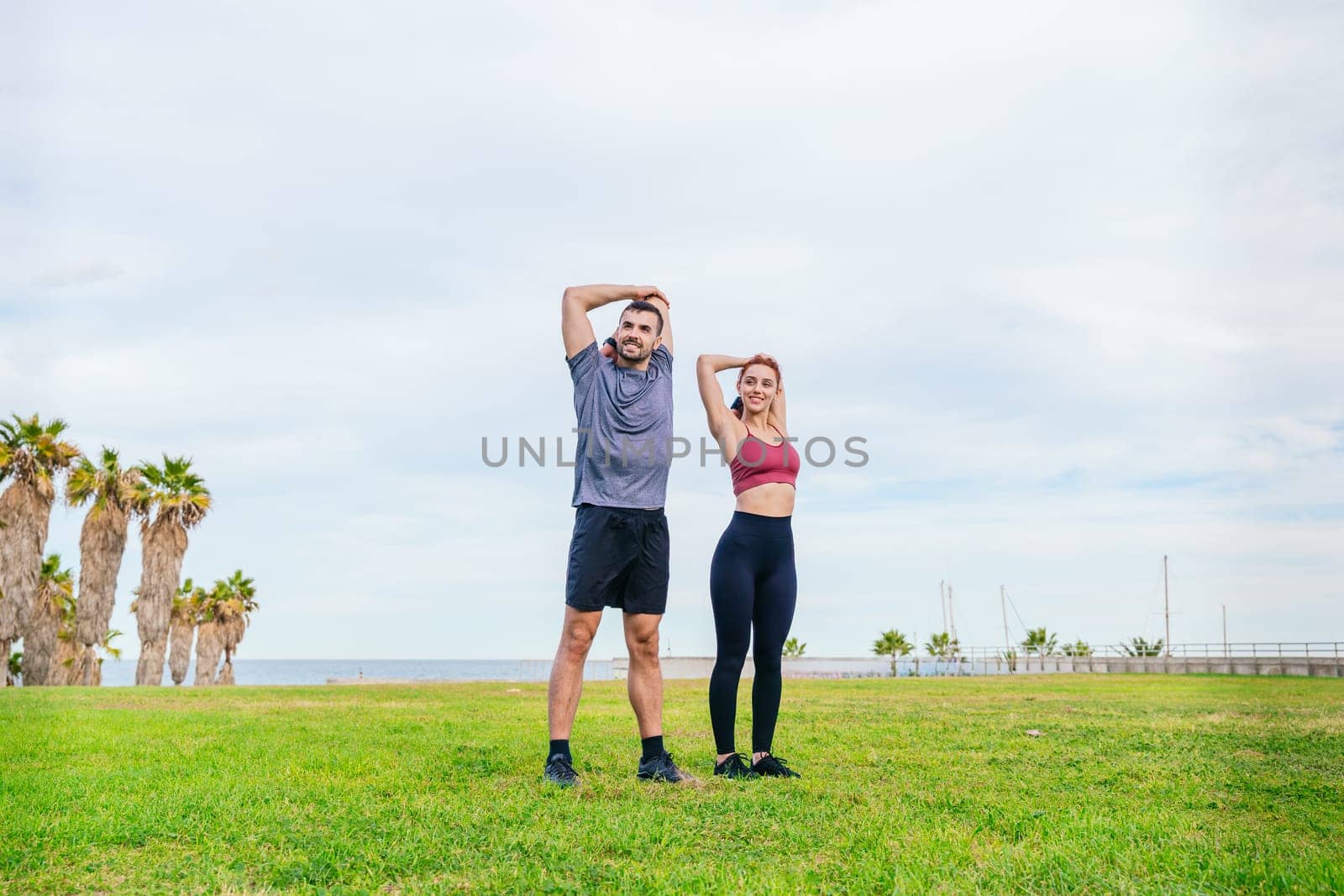 Young man and woman in sportswear stretching their arms in a green park near the coast. They are preparing for a workout, surrounded by palm trees and an open sky with fresh air.