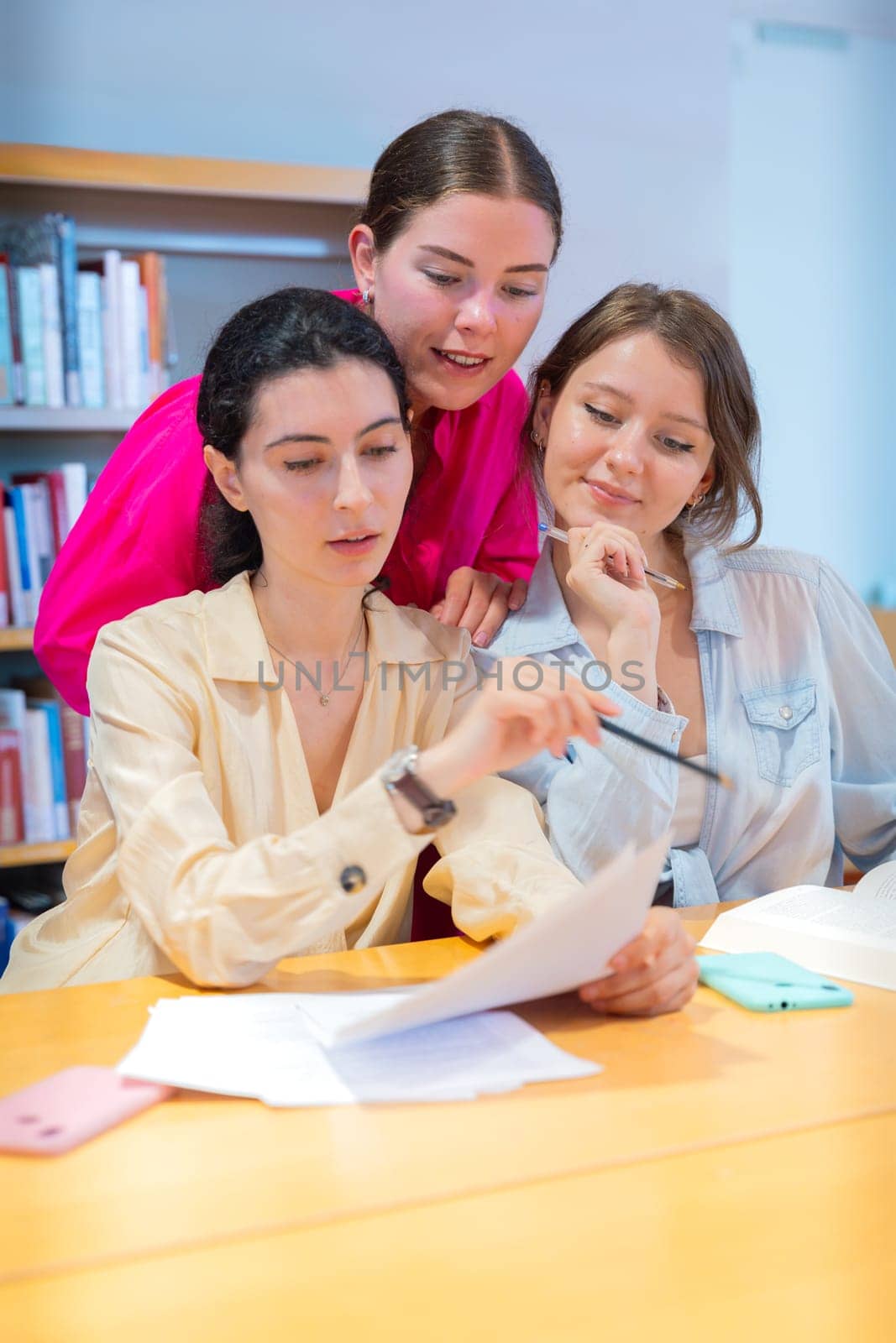 Three young women collaborating at a library table, studying and discussing documents. They are engaged in learning, taking notes, and exchanging ideas in a focused academic environment.