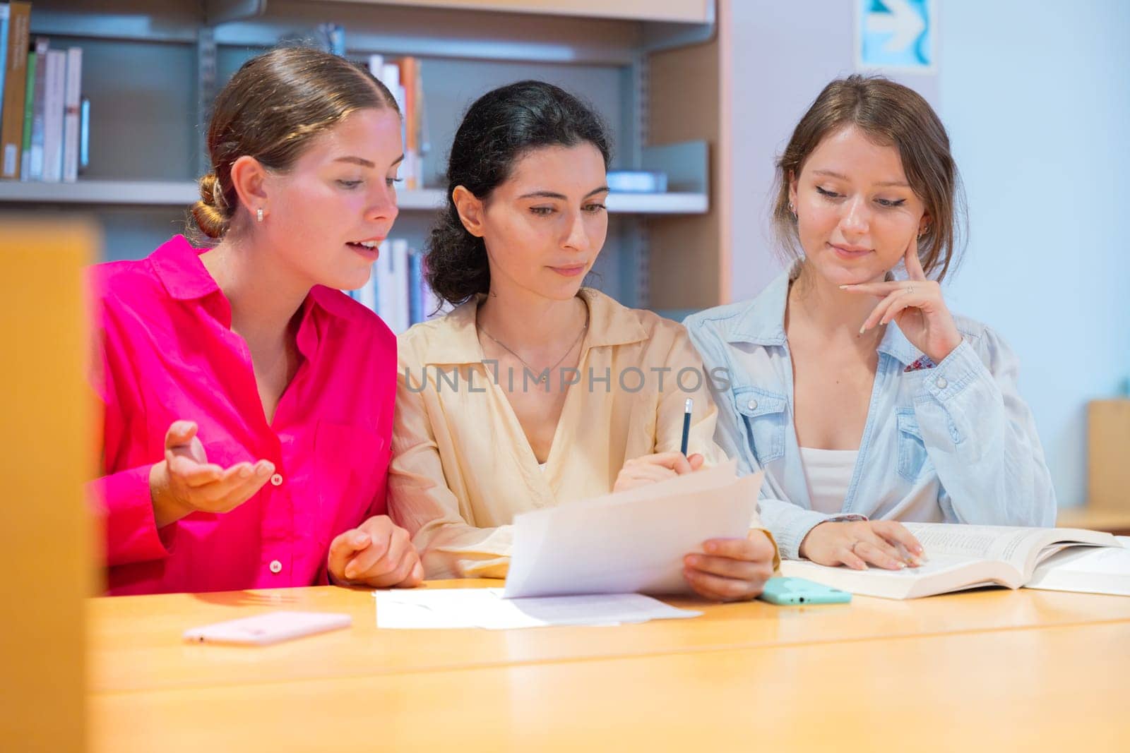 Three young women collaborating at a library table, studying and discussing documents. They are engaged in learning, taking notes, and exchanging ideas in a focused academic environment.
