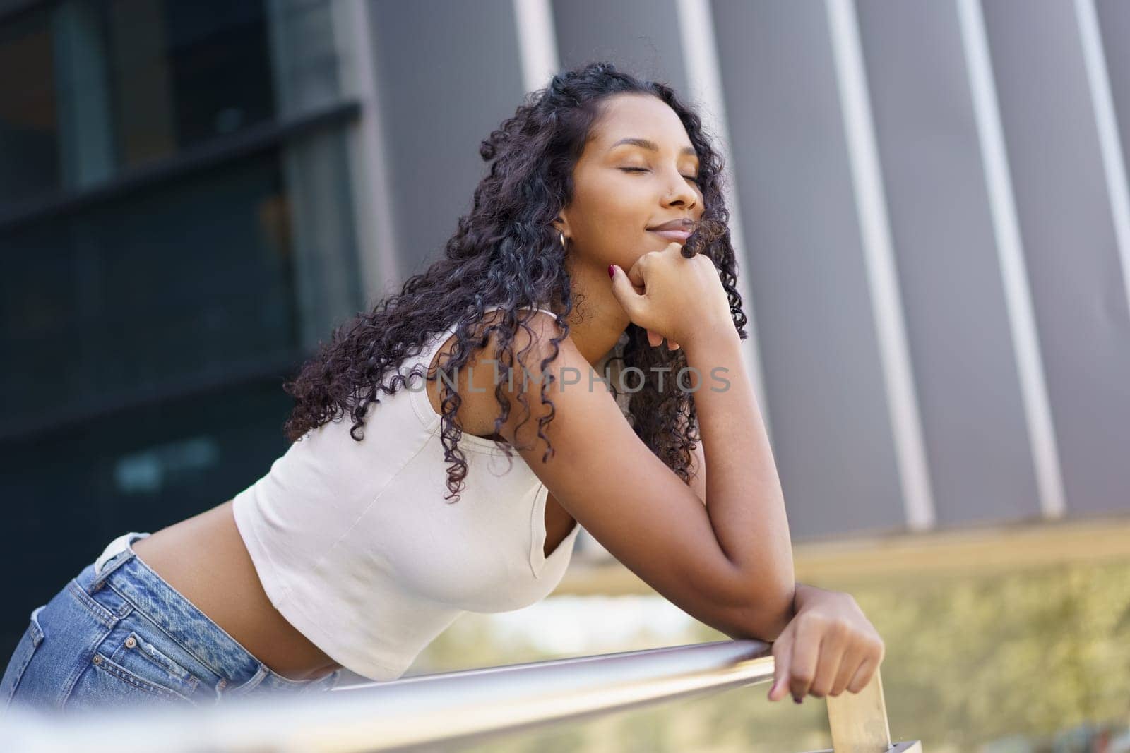 A young woman with curly hair leans thoughtfully against a decorative railing, embodying serene tranquility and stylish poise in a vibrant urban setting filled with life and energy