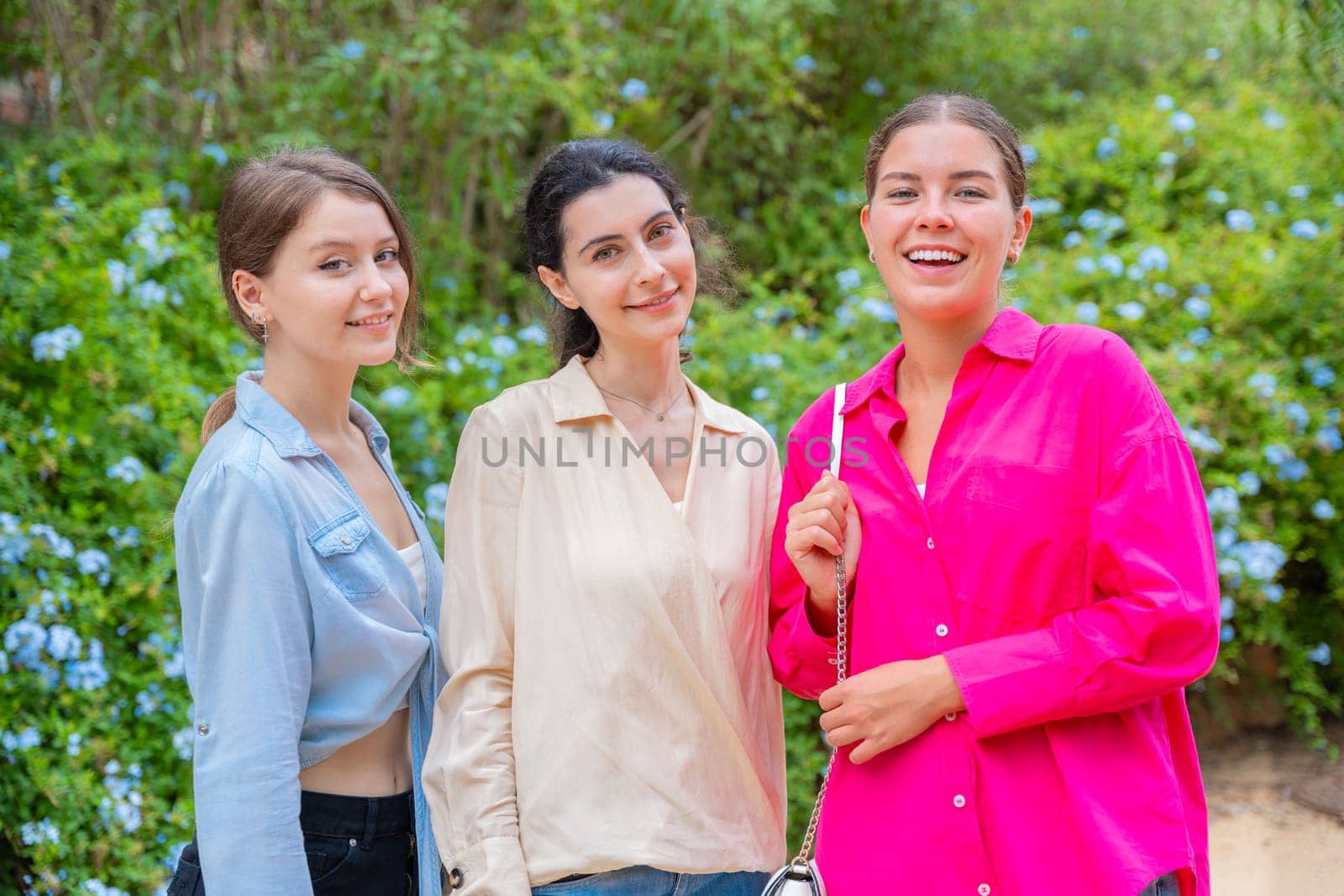 Three young women smiling and embracing each other while posing in a lush green park. They wear stylish casual outfits, expressing happiness, friendship, and joy in a natural outdoor setting.