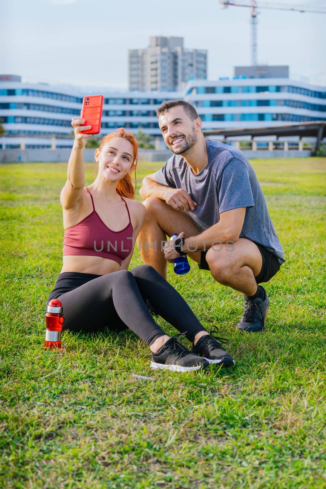 Smiling young man and woman sitting on the grass, taking a selfie after exercising. They are wearing sportswear and holding water bottles, enjoying a relaxed moment in an urban park.