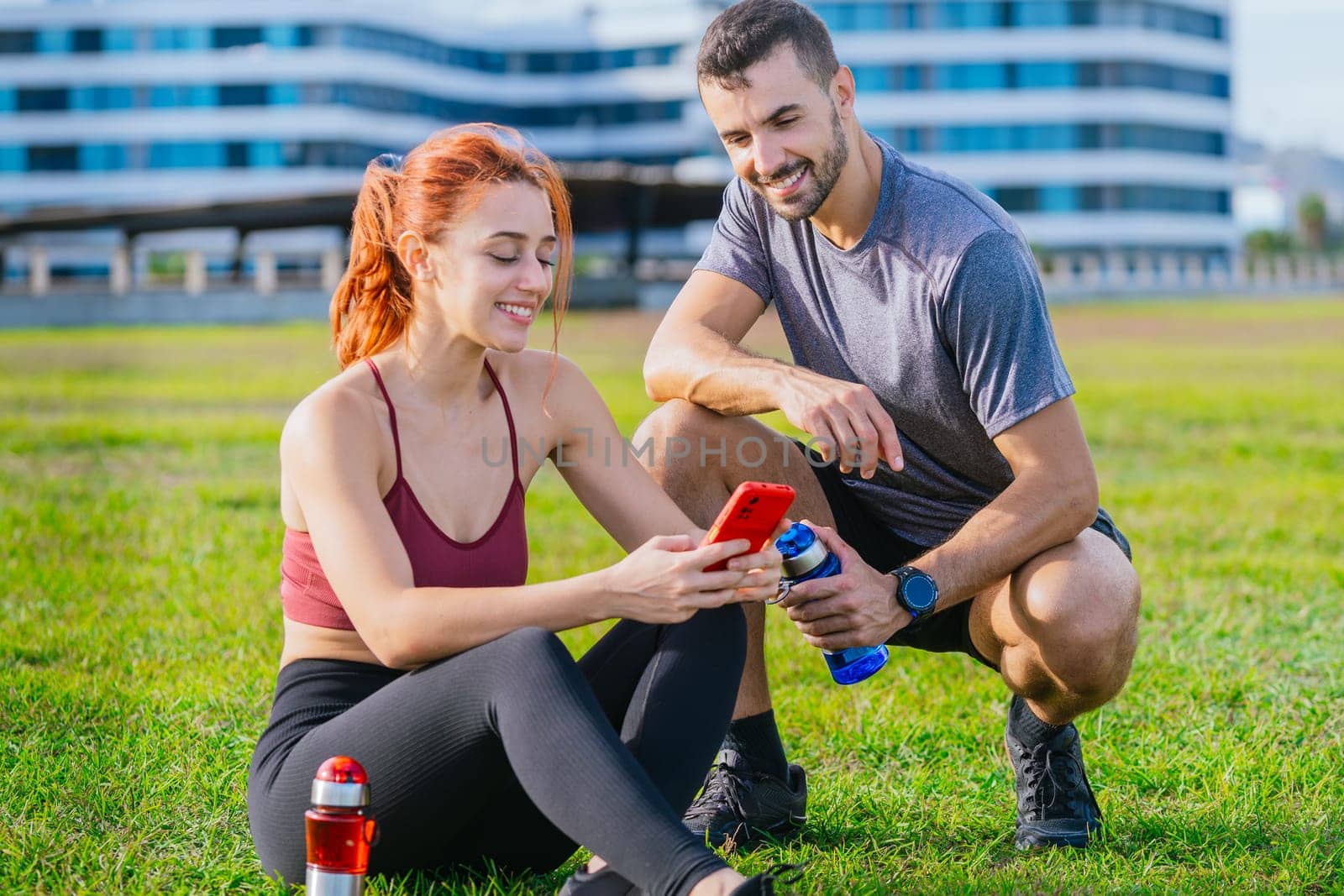 A young fit couple in sportswear relaxing after exercise in a green park. The woman is sitting on the grass using her smartphone, while the man crouches beside her with a water bottle, both smiling.