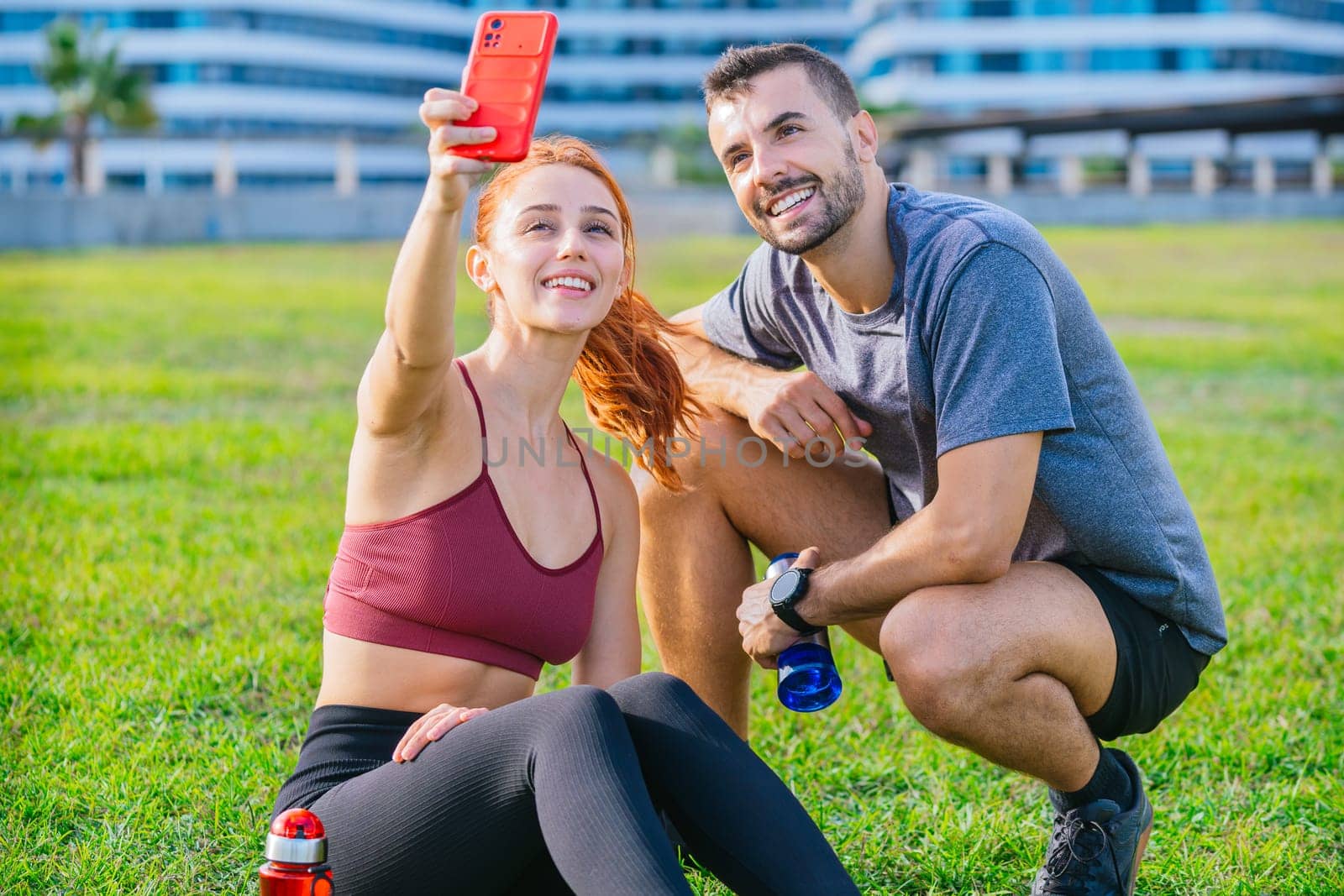 Smiling young man and woman sitting on the grass, taking a selfie after exercising. They are wearing sportswear and holding water bottles, enjoying a relaxed moment in an urban park.