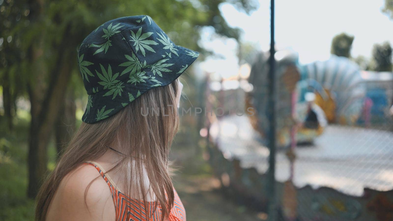 Young woman with marijuana leaf patterned hat looking at a carousel in a park during summer