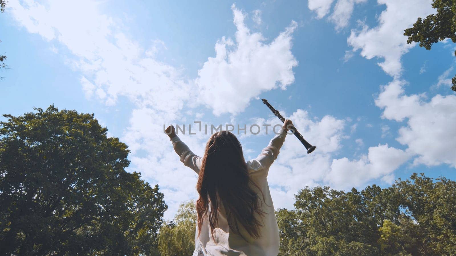 Young musician raising clarinet, arms extended victoriously after triumphant performance amid verdant park landscape, sunlight illuminating blue sky backdrop