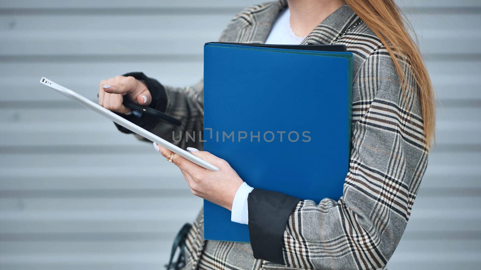 Professional businesswoman working on tablet, holding blue folder, pointing at screen while wearing white shirt and checkered jacket
