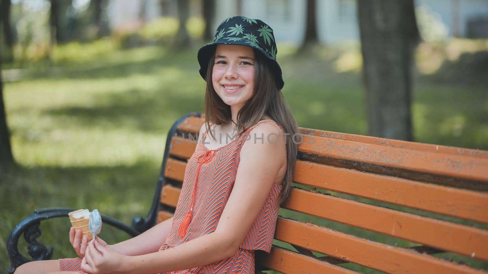 Smiling teenage girl wearing bucket hat sitting on park bench and eating ice cream cone in summer