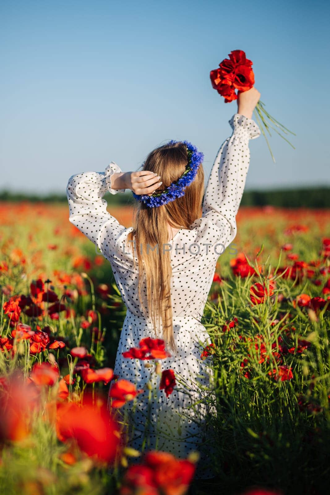 Blonde woman wearing white polka dot dress and flower crown holding red poppies in field enjoying summer