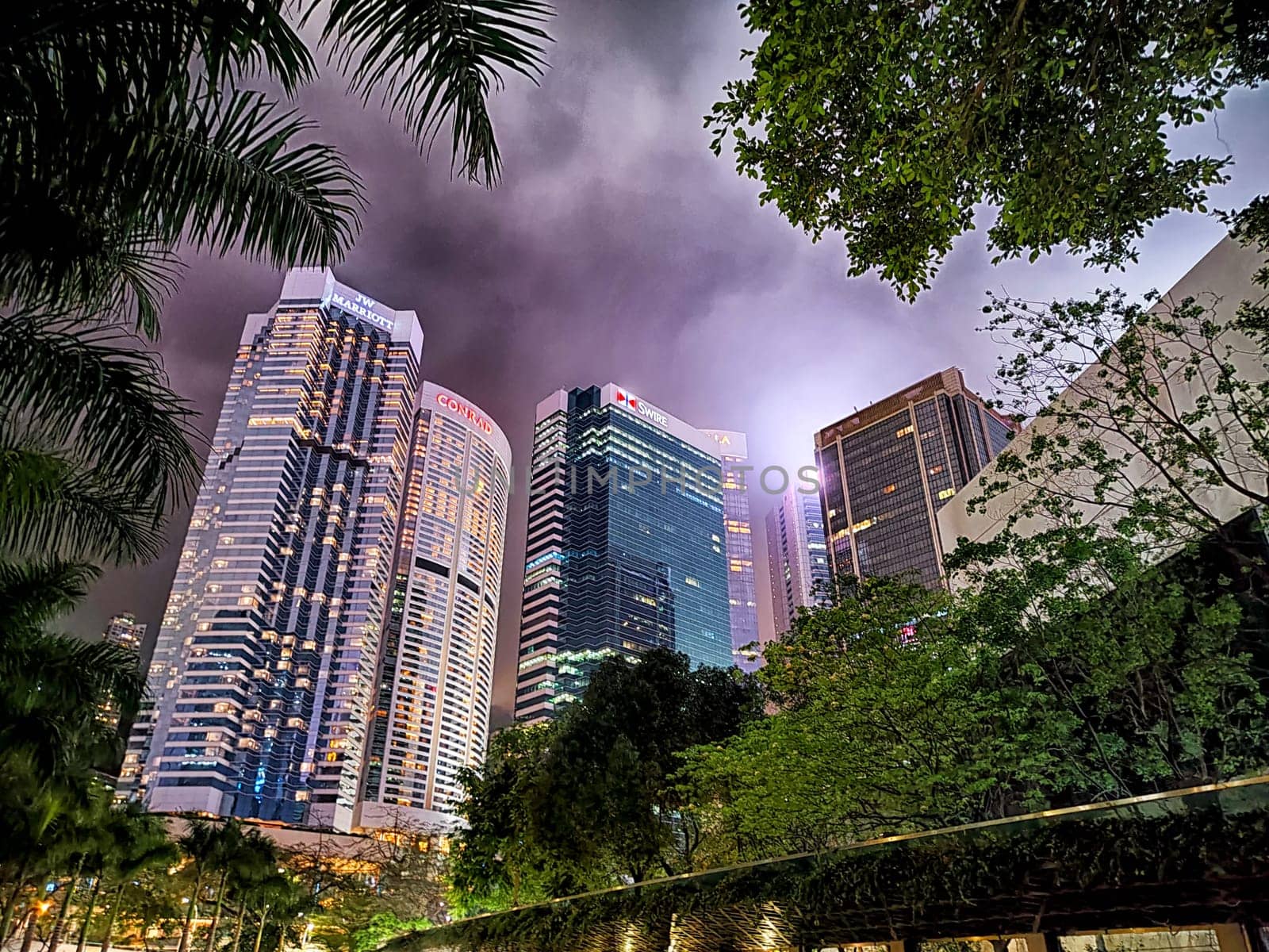 Hong Kong, China - April 06, 2024 - Tall skyscrapers in Hong Kong shine brightly against a moody sky, surrounded by lush greenery creating a vibrant urban atmosphere.