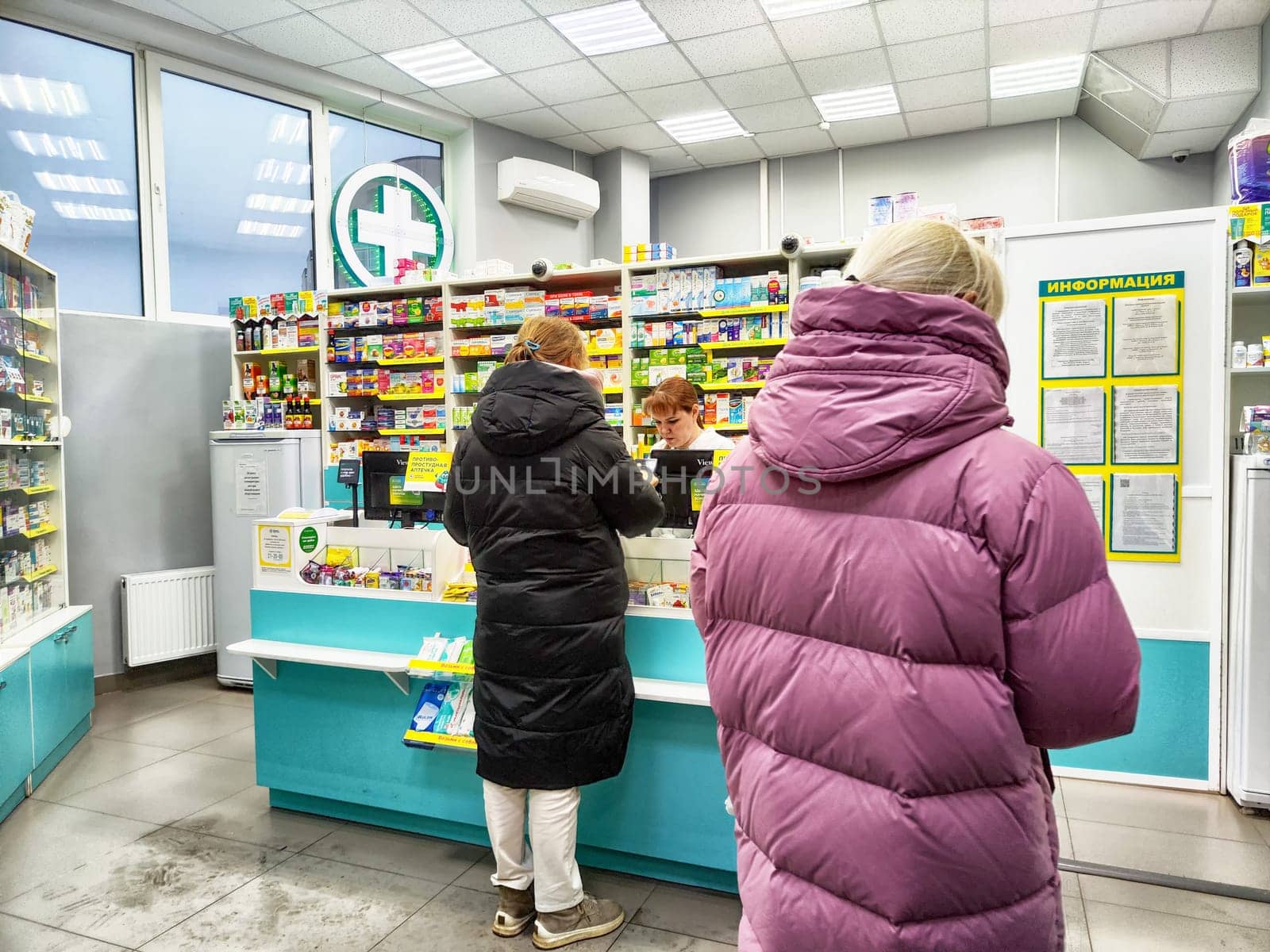 Kirov, Russia - December 28, 2024: Two customers stand at the pharmacy counter, browsing products while a staff member assists them with inquiries.