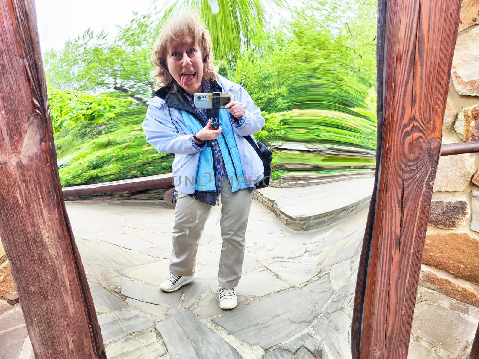 Blonde woman playfully poses for a selfie in a curved mirror, embracing the fun of nature in a park filled with greenery.