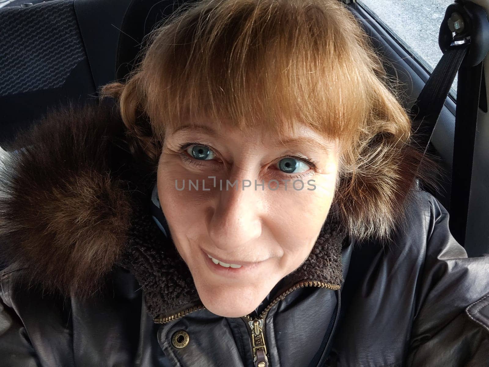 A blonde woman smiles at the camera from inside her car, enjoying a travel excursion on a beautiful day, sharing her adventures online.