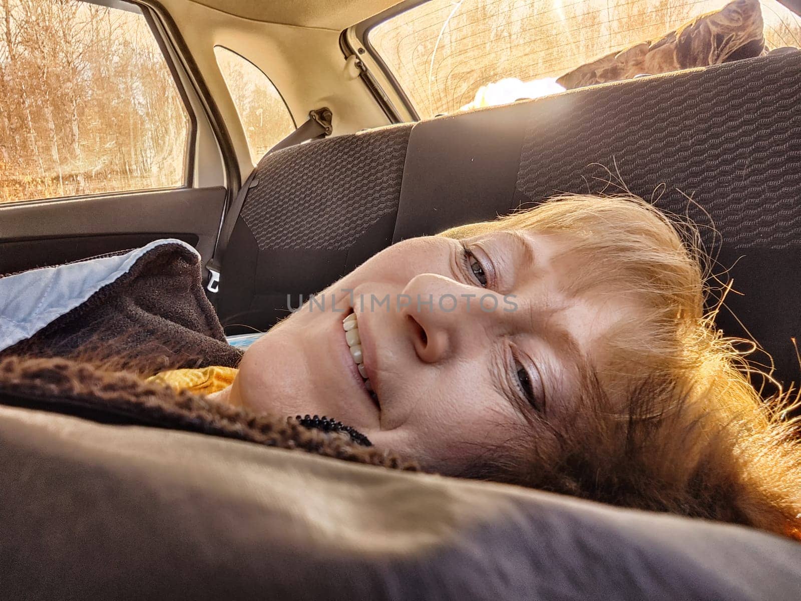 A middle-aged woman with blonde hair relaxes and naps in her car, feeling fatigued but content during her travels.
