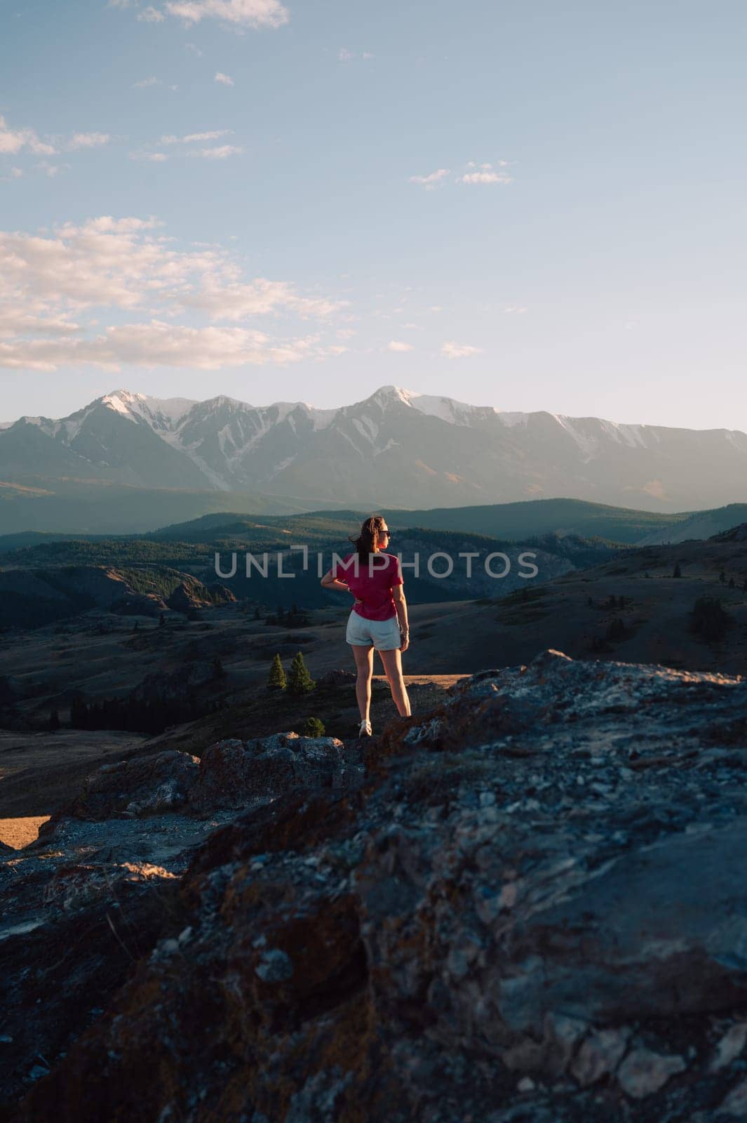 A woman in casual clothing stands on a rocky peak, gazing at scenic mountains and a vast valley under golden sunlight.